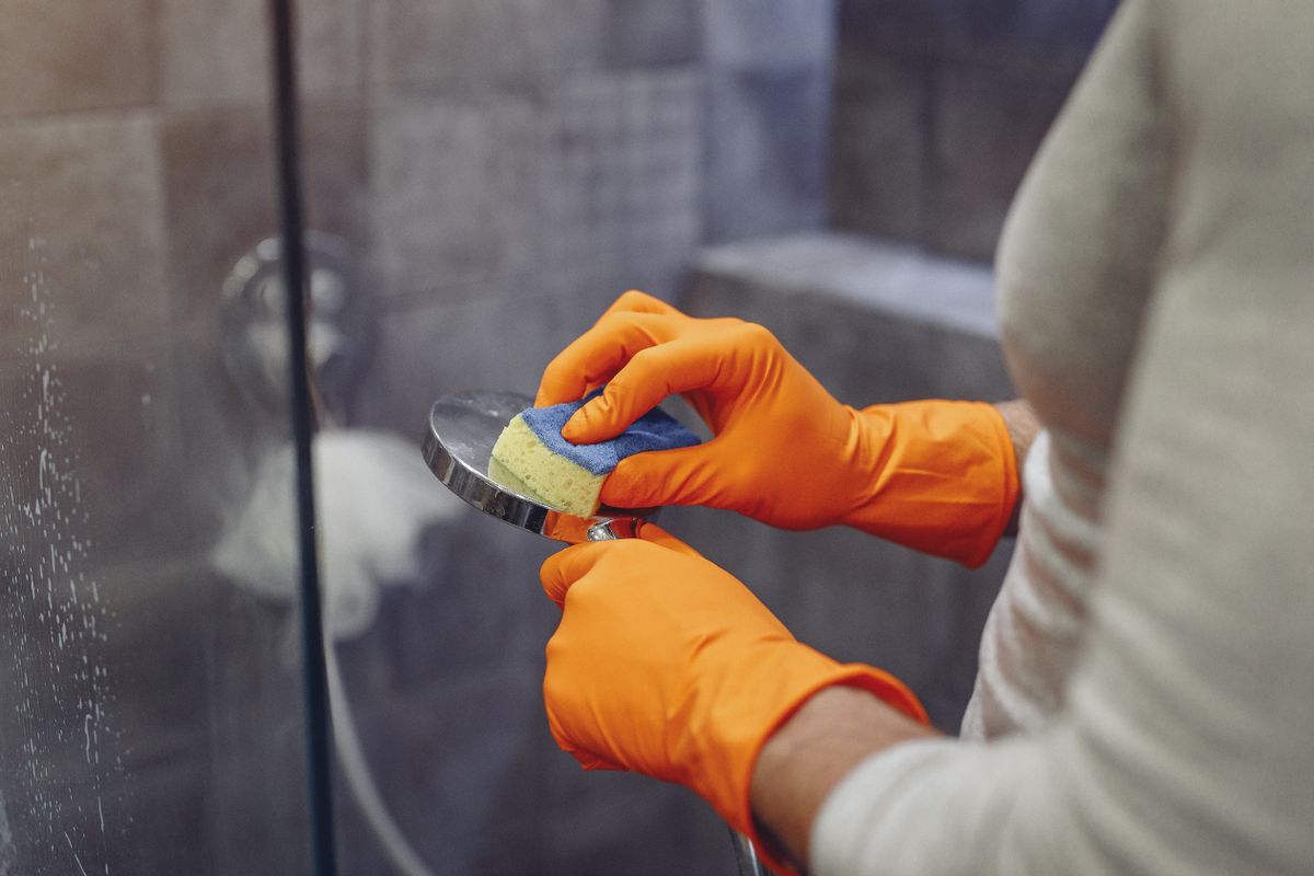 Gloved hands scrubbing a chrome shower head beside a glass shower door spotted with water marks