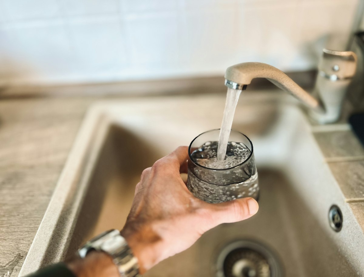 A hand filling a drinking glass with water from a kitchen sink faucet