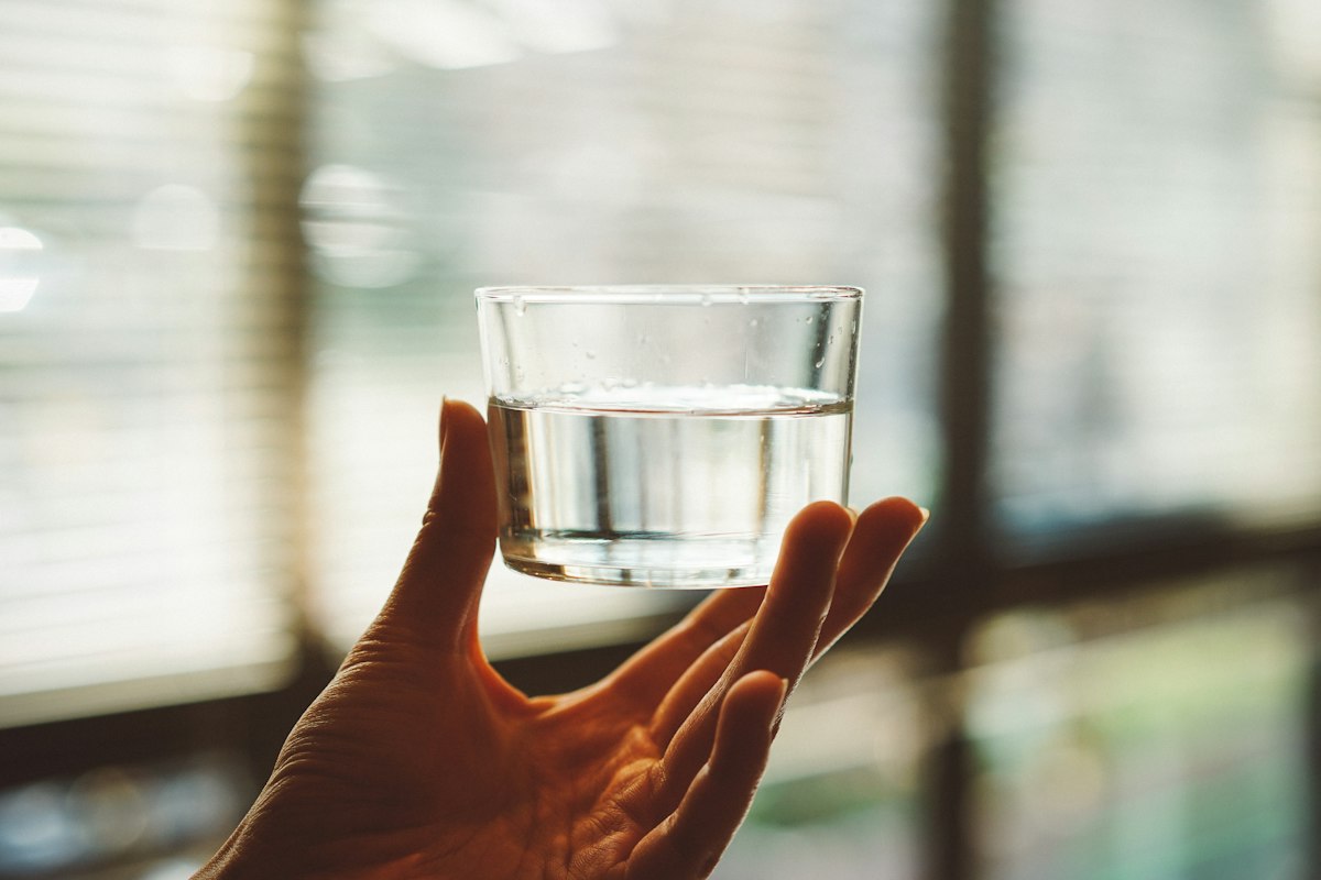 A hand holding a clear glass of water up to window light to check its clarity