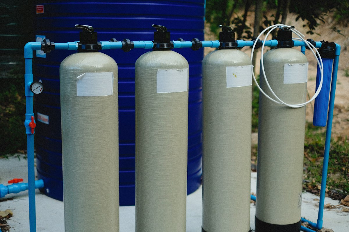 A row of four fiberglass filtration tanks with top-mounted control valves, joined by blue pipe beside a cartridge filter housing